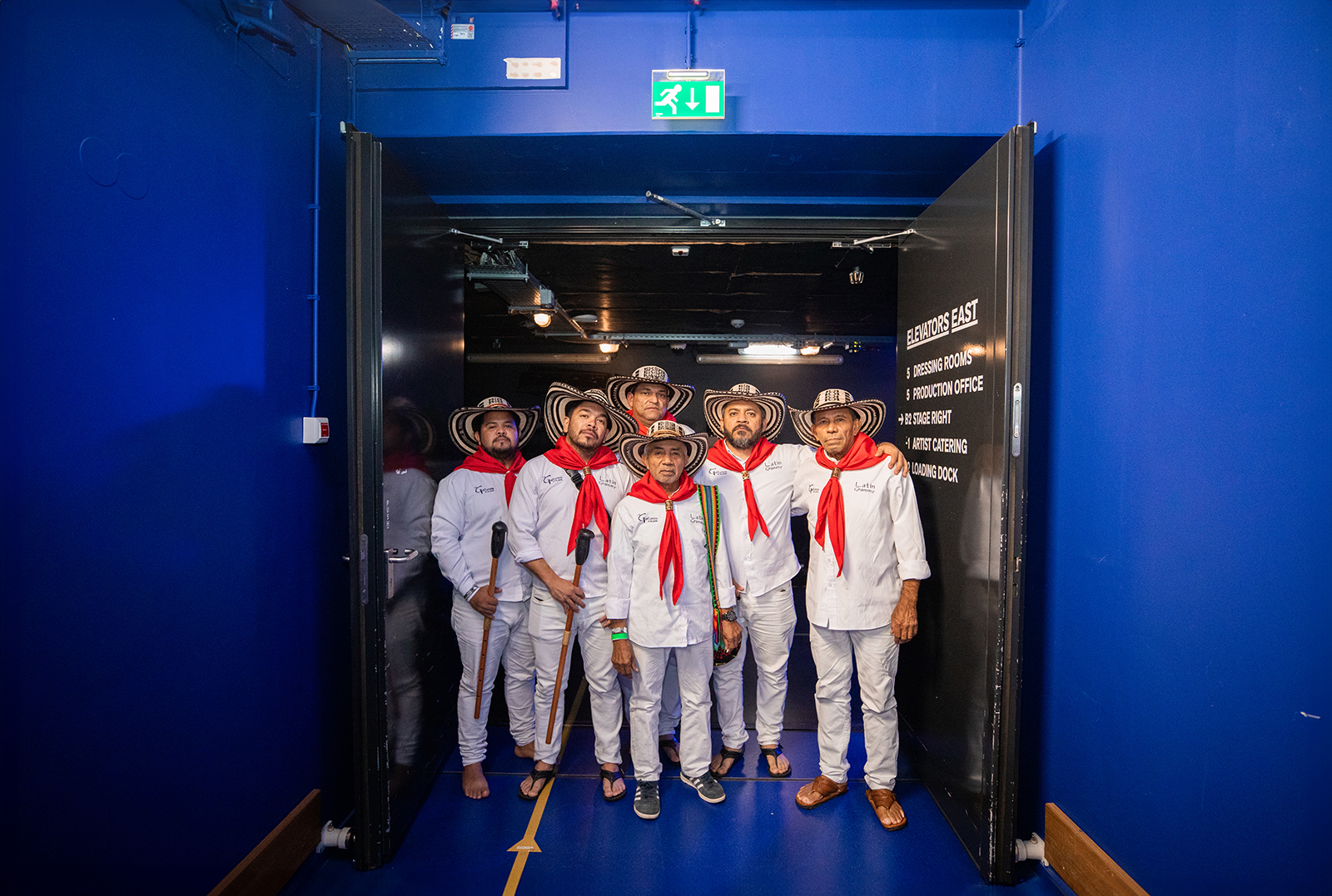 Photo of the members of Los Gaiteros de San Jacinto. They are standing in the doorway. They are all wearing white clothes, a red scarf and a hat.